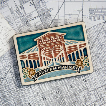 This Eastern Market Tile features the brick facade of the shed building. The words "Eastern Market" are written on a banner across the bottom of the tile with colorful flowers on either side.