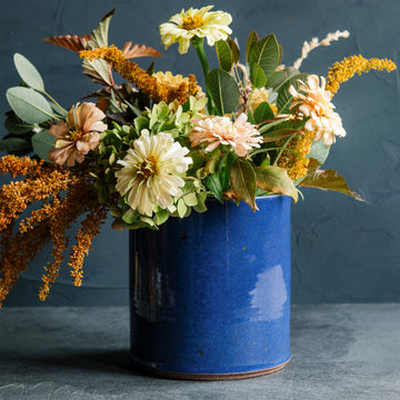 The crock is a cylindrical vessel with straight sides that has a wide opening and is as wide as it is tall. This Pewabic crock features the glossy, steel blue Ultramarine glaze.