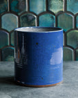 A close-up of the Crock in Ultramarine shows a smooth, glossy blue surface. Small brownish grey iron spots dapple the surface of the vase.