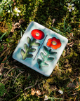 This handpainted tile in the Poppy Palette features bight red and deep orange petals and medium green stems. The background of the tile is a pale blue color.
