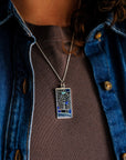 This brushed metal Detroit Map Pendant is rectangular with a line drawing of the areal view of a downtown Detroit street map. The word "Detroit" is written at the bottom of the design. Sections of the map are colored in varying shades of blue to delineate water and parks found around the downtown area.