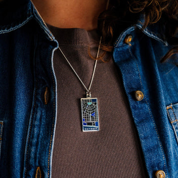 This brushed metal Detroit Map Pendant is rectangular with a line drawing of the areal view of a downtown Detroit street map. The word "Detroit" is written at the bottom of the design. Sections of the map are colored in varying shades of blue to delineate water and parks found around the downtown area.