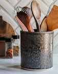 This Pewabic Crock sits on a white marble kitchen counter. The crock features the metallic gray textured Graphite glaze and holds wooden cooking utensils.