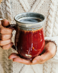 The smooth exterior of the glossy-red Winterberry mug shines in the sunlight while a woman in a warm sweater grasps it in her hands.