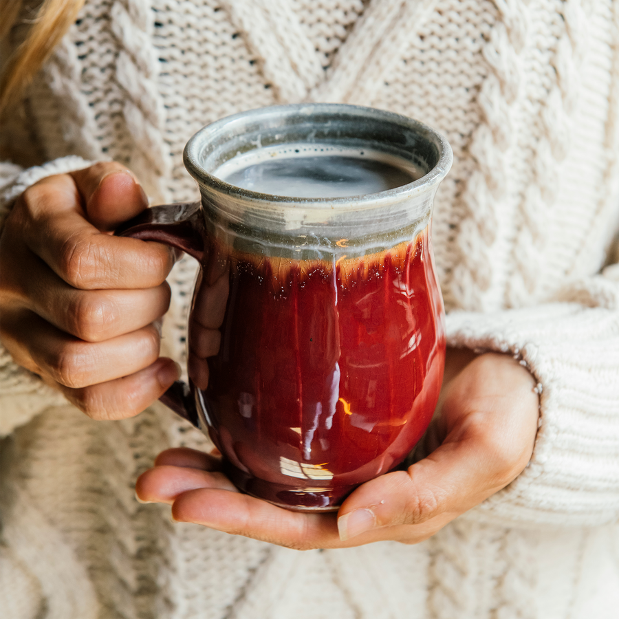 The smooth exterior of the glossy-red Winterberry mug shines in the sunlight while a woman in a warm sweater grasps it in her hands.