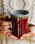 This Cafe Mug is being held by a woman in a cozy, creme-colored sweater. The base of the mug is resting in her hands.