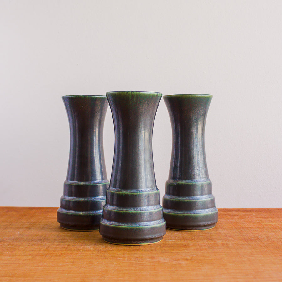 Three Step Vases in the Gun Metal Glaze sit together on a wood table. Each has slightly different amounts of the green lightening on the steps of the vase.