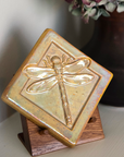 The Dragonfly Tile features a detailed dragonfly with wings outstretched. It is sitting horizontally on the square tile with a thick border around the edge. The pinkish gold metallic Blush Iridescent glaze features lots many variations. Depending on the lighting in the room, these pieces will pick up different hues.