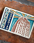 The Guardian Building Tile design features the top of the building on the right side of the tile with the word "Guardian" on the left. A large version of the Pewabic tiles found on the Guardian Building are framing the left side of the image. The tile is hand-painted in blues with red and yellow accents.