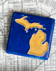 The Michigan Outline Tile features the outline of the state of Michigan with both peninsulas and its larger northern islands. This hand painted tile has a background of deep matte blue while the peninsulas are a maize gold color.