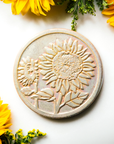 The Sunflower Trivet Tile features two sunflowers growing up into the circular tile with leaves and a bit of stalk in view. One sunflower is facing forward while the other is slightly smaller and facing to the left.