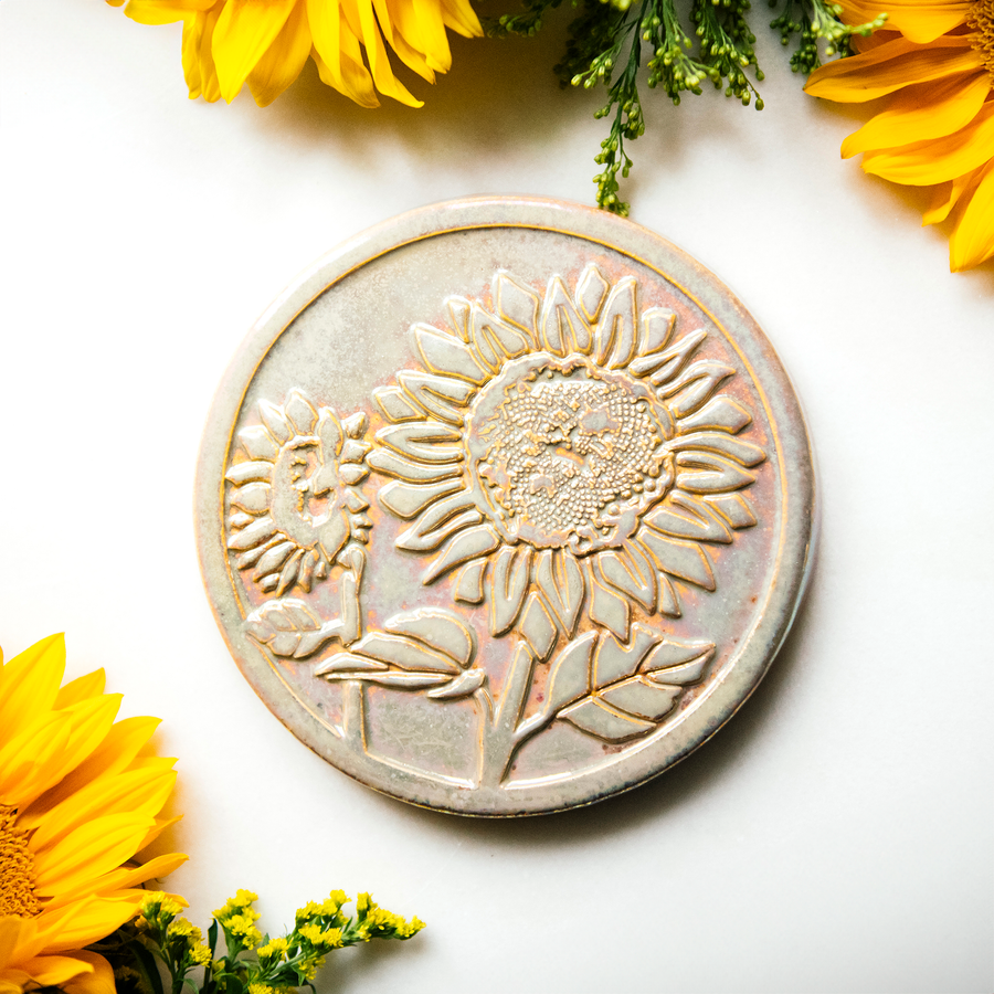 The Sunflower Trivet Tile features two sunflowers growing up into the circular tile with leaves and a bit of stalk in view. One sunflower is facing forward while the other is slightly smaller and facing to the left.