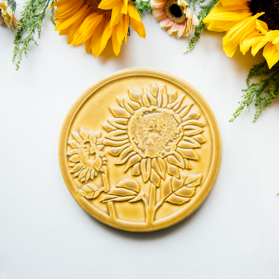 The Sunflower Trivet Tile features two sunflowers growing up into the circular tile with leaves and a bit of stalk in view. One sunflower is facing forward while the other is slightly smaller and facing to the left.