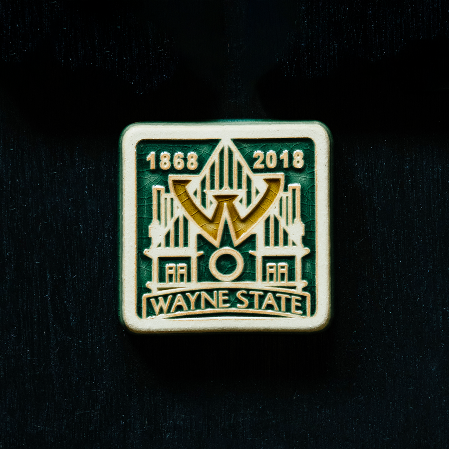 The Wayne State University tile is displayed on a map of Detroit. The tile boasts a prominent W in the center of the design that is hovering over a simple line drawing of the Old Main. On the bottom of the tile "Wayne State" is written in bold lettering and the dates 1868 and 2018 are displayed across the top- in celebration of Wayne State's 150 years as a learning institution. This whole design is depressed into a flat tile. The depressions hold glossy green and gold glaze.