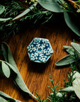 The Snowflake Pin rests in a medium wood board surrounded by winter greenery like juniper and sage.