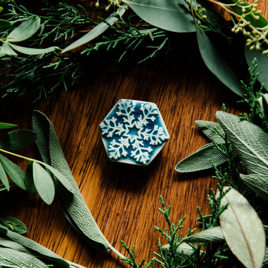 The Snowflake Pin rests in a medium wood board surrounded by winter greenery like juniper and sage.