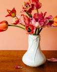 Our Snowdrop Vase in a white, slightly iron-speckled, "Birch" glaze rests on a dark wooden table. The vase is filled with bright-red and pink tulip varieties. The wall behind the vase is a peachy, springy, pink.