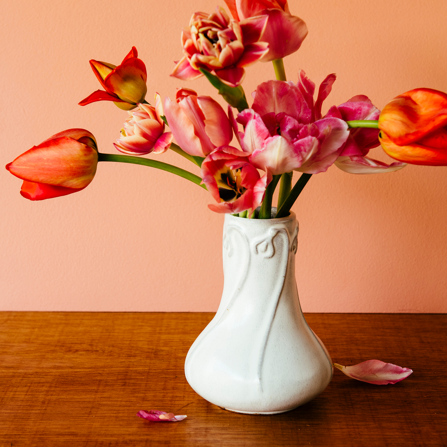 Our Snowdrop Vase in a white, slightly iron-speckled, "Birch" glaze rests on a dark wooden table. The vase is filled with bright-red and pink tulip varieties. The wall behind the vase is a peachy, springy, pink.