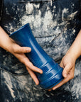 Two hands hold a Leleand Blue Step Vase. The person holding the piece is wearing a worn and dusty denim apron.