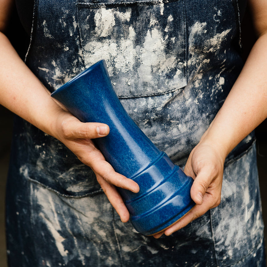 Two hands hold a Leleand Blue Step Vase. The person holding the piece is wearing a worn and dusty denim apron.