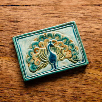 The Peacock Tile features a peacock facing straight ahead with its feathers fanned out showing off its colorful plumes. In the bottom right corner of the tile are the letters "M C S" in black writing. This is the only tile on which Pewabic's co-founder Mary Chase Stratton included a signature. The hand painted tile features bright blues, greens and beiges.