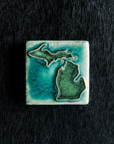 This hand painted tile has a matte turquoise background with matte earthy green peninsulas.