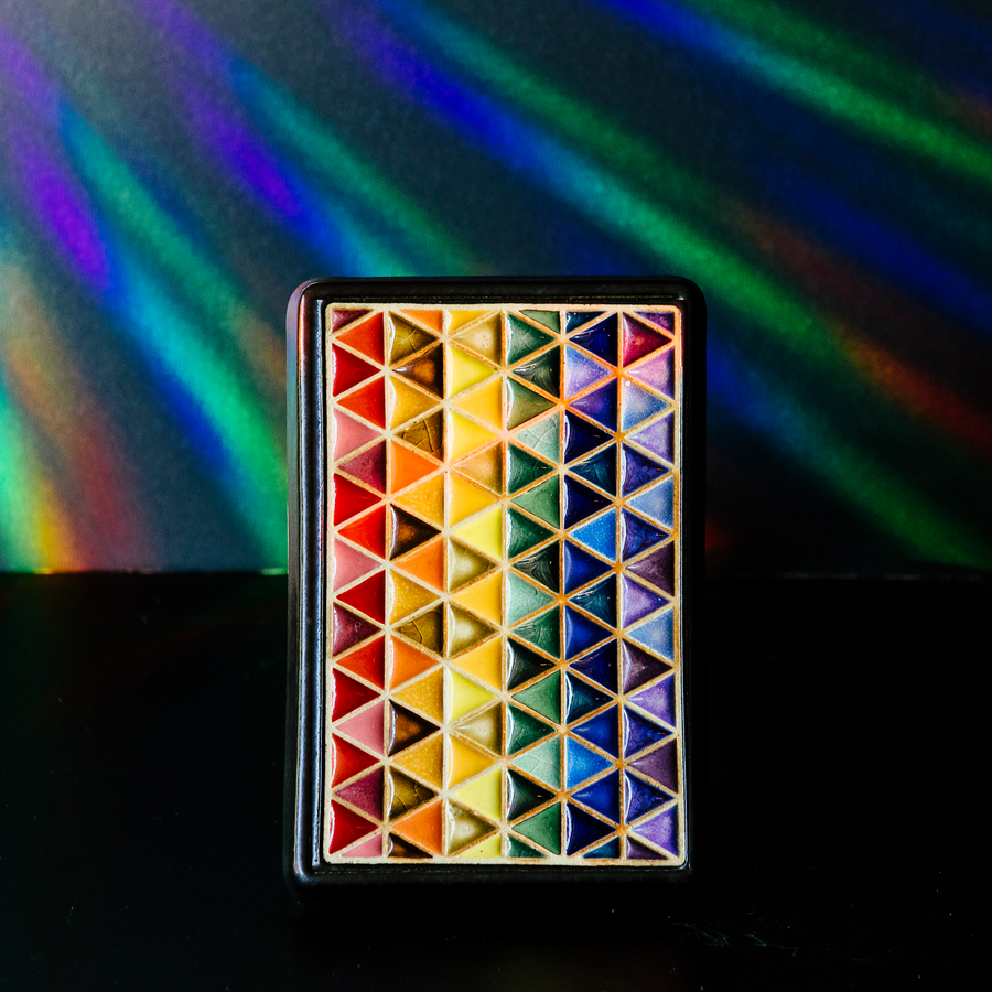 The 4x6 Pride Tile features lines of interlocking triangles, the colors on the triangles start at red and end in indigo- creating the rainbow Pride flag. The bright pattern is contained in a black border.