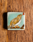 This Bird with Berries Tile depicts a brown bird sitting on a branch with red berries in its beak. This tile is hand-painted with brown, blue and red glazes.