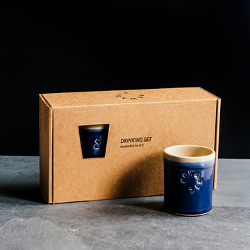 Box set of two glossy, dark-blue Midnight Rocks Cups. The box reads "Drinking Set - Handcrafted Set of 2." One of the cups is outside of the box to showcase its glaze surface against a black backdrop and slate foreground. 