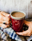 This Cafe Mug is being held by a woman in a cozy sweater with a blanket on her lap.