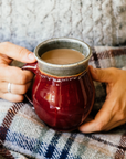 The smooth exterior of the mug shines in the sunlight while a woman in a warm sweater grasps it in her hands.